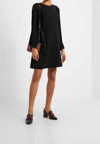 Schwarzes Kleid mit plissierten langen Ärmeln, rundem Halsausschnitt und einem ausgestellten Rock, kombiniert mit einer kleinen Tasche im rot-schwarzen Leopardenmuster und hochhackigen Schuhen.
