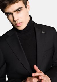 Czarna marynarka smokingowa o dopasowanym kroju, zestawiona z czarnym golfem. Warto zwrócić uwagę na subtelnym logo na klapie.