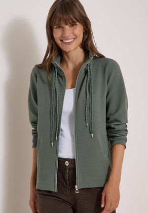 Donna sorridente che indossa una giacca verde con cerniera sopra una maglietta bianca e pantaloni marroni, in piedi davanti a uno sfondo chiaro e uniforme.