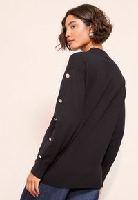 Pulover negru din tricot, cu o croială lejeră, având mâneci lungi și butoane decorative argintii pe latura exterioară a mânecilor.
