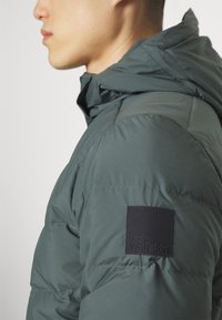 Grüne gepolsterte Jacke mit Kapuze, ausgestattet mit einem strukturierten schwarzen Logo-Patch am Ärmel, das ein gestepptes Design hervorhebt.