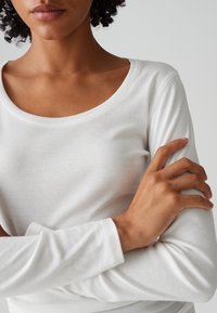 Frau mit mittlerem Hautton, trägt ein weißes Langarmshirt, die Arme verschränkt, vor einem einfarbigen hellgrauen Hintergrund.