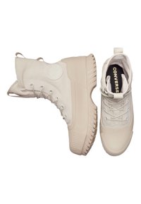 Zapatillas altas blancas con suela de goma texturizada, parte delantera con cordones y parche del logo de Converse. Materiales sintéticos y de lona suaves.