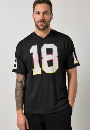 Mann mit Bart und tätowiertem rechten Arm, der ein schwarzes Sporttrikot mit großer weißer Nummer 18 trägt, rot und gelb umrandet auf Vorderseite und Ärmeln.