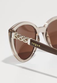 Ochelari de soare tip cat-eye cu rame transparente de culoare căprui deschis, lentile de culoare căprui închis și un detaliu din lant auriu pe brațe. Logo "Vogue" în galben.