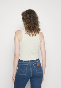 Top de alças largas em verde claro com nervuras, combinado com jeans ajustados em azul escuro com um remendo de couro castanho e costura distinta nos bolsos traseiros.