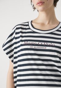 Pasiasta bawełniana koszulka w kolorze granatowym i białym, z krótkimi rękawami, z okrągłym dekoltem i grafiką z przodu z nazwą marki oraz rokiem założenia.