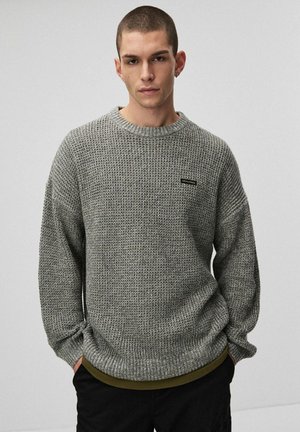 Szary sweter z dzianiny o teksturowanym wzorze, okrągłym dekolcie i subtelną naszywką z logo nad dolnym brzegiem. Długie, lekko oversize'owe rękawy.