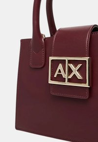 Borsa a mano in pelle sintetica bordeaux con forma strutturata, hardware con logo dorato e manici corti. Presenta una pattina con chiusura a ribalta e dettagli di cucitura.