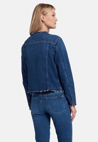Giacca di denim blu scuro con orlo e polsini sfrangiati, caratterizzata da un taglio classico e due tasche posteriori; mostrata da un angolo laterale.