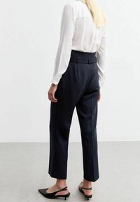 Pantalones oscuros de navy ajustados con una cintura alta, pierna recta, bolsillos laterales y un sutil detalle en el cinturón, combinados con una blusa blanca de manga larga.