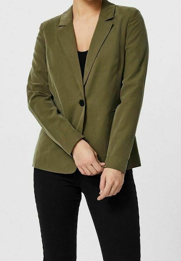 Femme portant un blazer vert olive à un seul bouton sur un haut noir et un pantalon noir, debout les mains jointes devant elle.