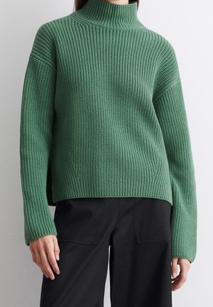 Personne portant un pull à col roulé vert en grosse maille avec manches longues, associé à un pantalon noir, debout contre un mur clair uni.