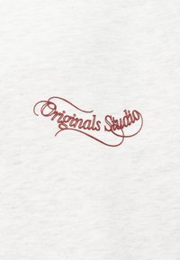 Λευκό ύφασμα με κόκκινο κεντητό κείμενο «Originals Studio» σε ρέον στυλ γραφής. Η υφή είναι απαλή και ελαφριά.