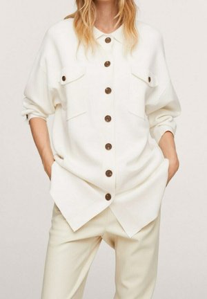 Kobieta ubrana w oversize'ową białą koszulę na guziki z brązowymi guzikami i dopasowane białe spodnie, z rękami w kieszeniach.