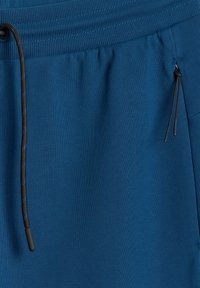 Pantalon de survêtement bleu avec taille côtelée, cordon noir et poche latérale zippée munie de tirettes noires.