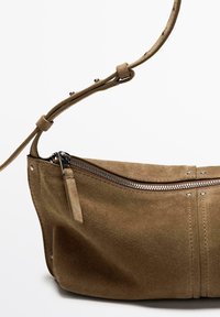 Sac à main en daim marron avec fermeture éclair argentée et sangle ajustable sur fond blanc uni.