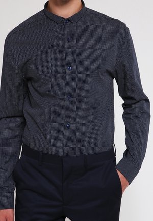 Man draagt een aansluitend donkerblauw overhemd met kleine witte stipjes en donkerblauwe broek, hand in de zak.