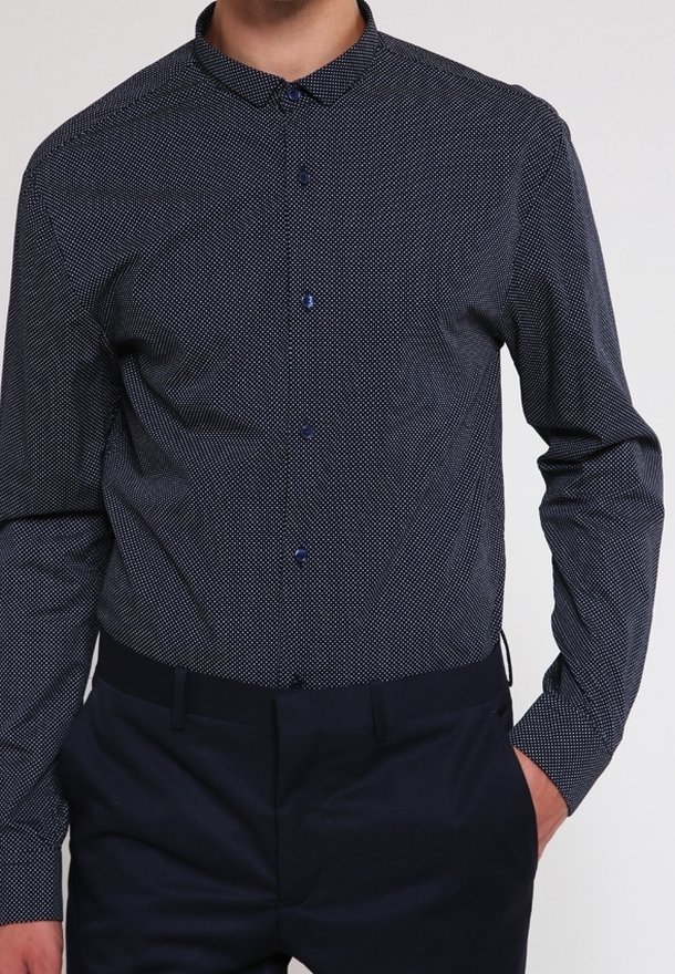 Homme portant une chemise ajustée bleu foncé avec de petits points blancs et un pantalon bleu foncé, main dans la poche.