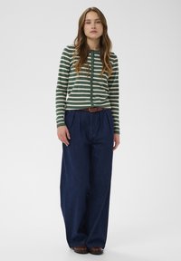 Sieviete stāv ar garām brūnām matām, ģērbta zaļi un baltos svītrainos pogātajos džemperī, platās tumši zilās biksēs, ar brūnu jostu un brūnām kurpēm.