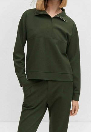 Femme portant un pull à manches longues vert foncé avec un court zip et un pantalon assorti, debout avec une main dans la poche.