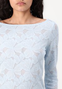 Light blue lace top featuring floral patterns, long sleeves, and a wide neckline, with a sheer texture and layered design.