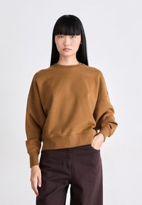 Felpa marrone realizzata in un tessuto morbido, con scollo rotondo e vestibilità rilassata, dotata di maniche a raglan e orlo a costine. Pantaloni scuri la completano.