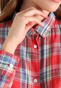 Camisa de cuadros roja con patrón de cuadros en azul, verde y negro. Presenta un cuello abotonado y botones blancos en la parte delantera y los puños.