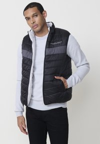 Schwarze und graue Steppweste mit gep quilteter Textur, mit Reißverschluss vorne und seitlichen Taschen. Getragen über einem hellgrauen Pullover.