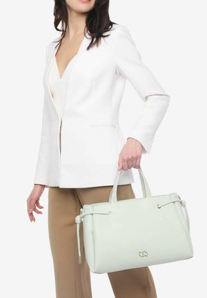 Mujer con blazer blanco y pantalones beiges sosteniendo un bolso estructurado de color verde pastel claro con doble asa y nudos laterales.
