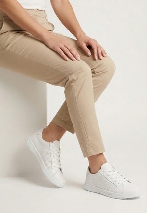 Person sitzt mit Händen auf beige Hose ruhend, trägt weiße perforierte Schnürsneakers vor hellem Hintergrund.