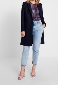 Abrigo largo azul marino sobre una camiseta estampada, jeans rectos de un azul claro y sandalias de tacón rosa con tiras, exhibiendo diversas texturas y colores.