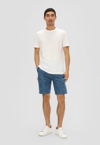 Witte katoenen t-shirt, blauwe denim cargo shorts met zijzakken, en witte sneakers; model staat voor een neutrale achtergrond.