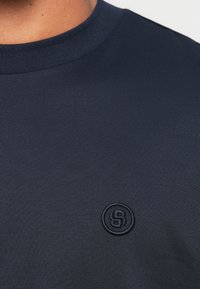 Granatowy t-shirt z okrągłym dekoltem wykonany z gładkiej tkaniny, z subtelnie wyszywanym logo na piersi i teksturowanym prążkowanym wykończeniem przy dekolcie.