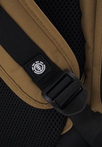 Primer plano de una correa de mochila marrón y negra con una hebilla de plástico negro y un logo de marca circular blanco sobre tela negra.