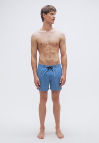 Μπλε μαγιό βερμούδα με ελαστική μέση, μαύρο κορδόνι και λεία υφή. Φοριέται από έναν άνδρα μοντέλο που στέκεται ξυπόλυτος, κοιτάζοντας μπροστά.