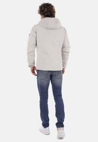 Chaqueta con capucha de color gris claro y textura suave, con cierre de cremallera y bolsillos laterales, llevada sobre unos vaqueros de mezclilla azul y zapatillas blancas.