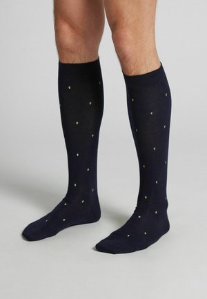 Ben iført høje sorte sokker med små grønne kaktusmønstre, stående på en lysegrå gulv mod en ensfarvet baggrund.