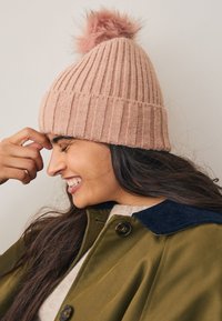 Rózsaszín kötött sapka bolyhos bojttal a tetején, függőleges bordázattal és szoros illeszkedéssel, zöld dzsekivel és tengerészkék gallérral együtt viselve.