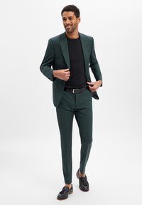 Grüner Anzug mit maßgeschneiderter Passform und feiner Textur, kombiniert mit einem schwarzen T-Shirt und schwarzen Loafers mit Quastenakzenten.