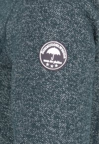 Tmavě zelená skvrnitá látka s kulatou nášivkou, na které je bílý deštník a německý text "Schmuddelwudda wenn dit plästert".