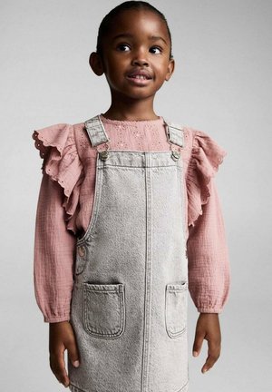 Graues Denim-Latzkleid mit zwei Fronttaschen, kombiniert mit einer pinkfarbenen Bluse mit Rüschensäumen und Lochmuster.