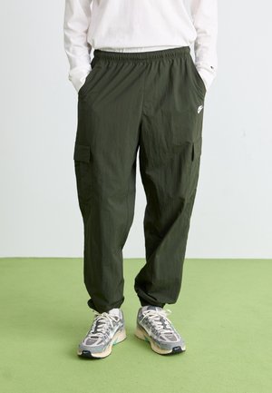 Personne portant un pantalon cargo vert olive avec taille élastique et des baskets blanches et grises debout sur un sol vert.