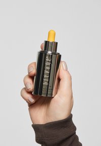 Metallisk silverserumburk med en gul pipettkork; har präglad text och en slät, reflekterande yta. Hand som håller i burken.