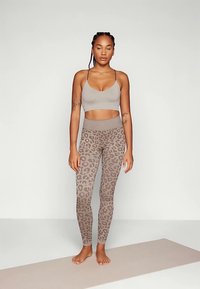 Nő mezítláb áll egy jógamatracra, szemét lehunyva, bézs sportmelltartót és leopárdmintás leggingset visel.