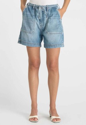 Vrouw draagt lichtblauwe denim shorts met grote voorzakken en een trekkoord in de taille, gecombineerd met witte sandalen met hak.