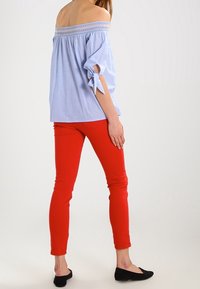 Hellblaues Off-Shoulder-Oberteil mit elastischem Saum und gebundenen Ärmeln, kombiniert mit figurbetonten roten Hosen und schwarzen Ballerinas. Glatte Stoffqualität.