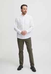 Mann står med hendene foldet, iført en hvit skjorte med knapper, olivengrønne bukser og svarte sko, mot en enkel lys bakgrunn.