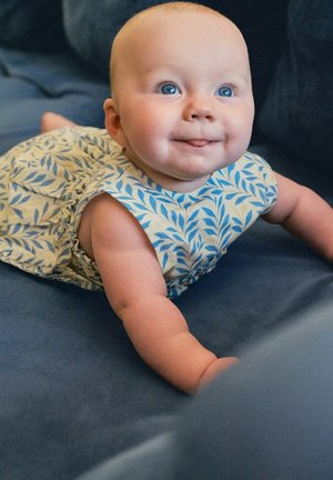 Bambino con occhi azzurri in un vestito azzurro e bianco con motivo a foglie, sdraiato su una superficie blu scuro, sorridente e che guarda leggermente verso l'alto.