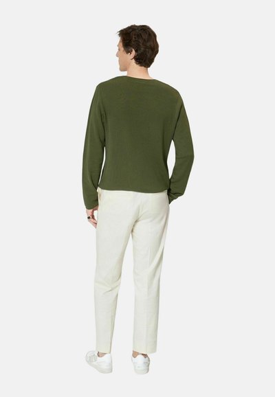 Hombre con camisa de manga larga verde oliva, pantalones blancos y zapatillas blancas de pie de espaldas contra un fondo liso.